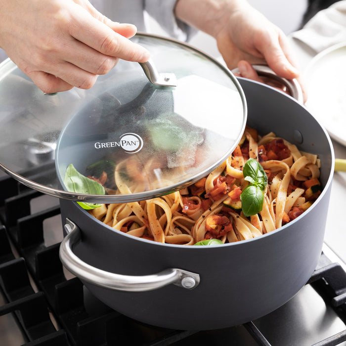 Hand lifting lid of gray ceramic 5-quart stockpot with fettuccine, tomato sauce, and basil inside.