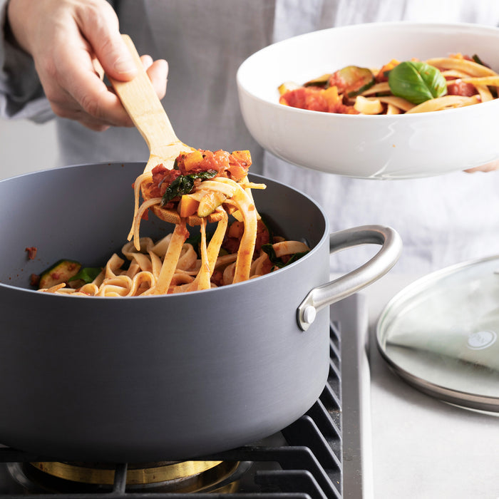 Vegetable pasta being served from a green ceramic nonstick stockpot with glass lid into a white bowl on a stovetop.