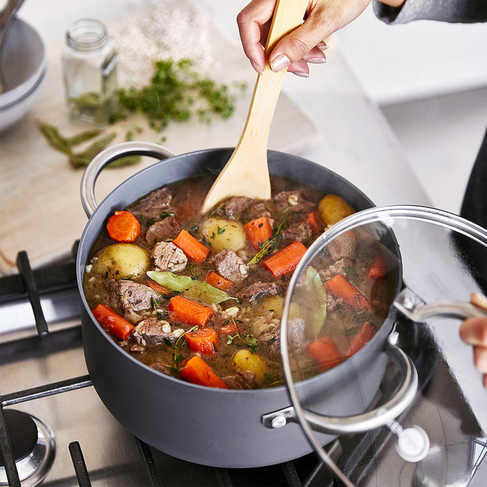 Beef stew with potatoes and carrots being stirred in a ceramic nonstick stockpot, lid held to the side.
