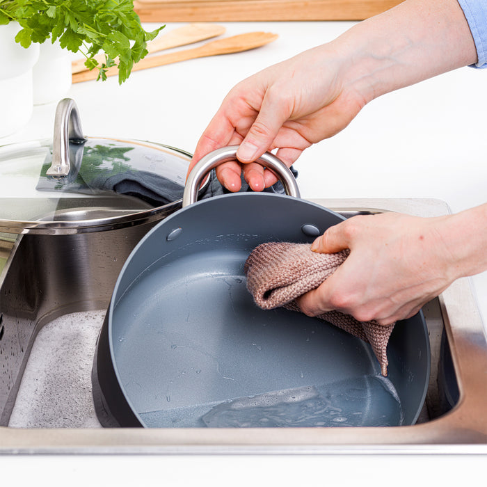 Person washing a nonstick pan at the sink next to a green 5-quart ceramic stockpot with lid; herbs in the background.