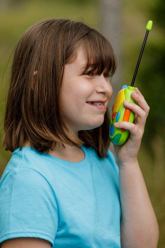 A smiling young girl with brown hair and braces, in a light blue shirt, uses the Outdoor Discovery Walkie Talkie by Outdoor Discovery, speaking into it while outdoors.