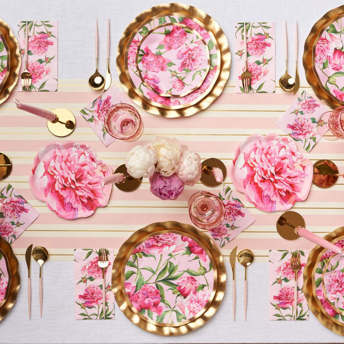 A table is set with pink and gold partyware, gold cutlery, pink glasses, candles, and Sophistiplate's Cocktail Napkin Pink Peonies. Large pink peonies decorate the center atop a striped cream and pink table runner.