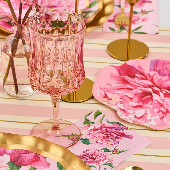 A pink and gold table setting with Sophistiplate's Cocktail Napkin Pink Peonies, pink crystal glass, floral plates, gold utensils, and a vase of pink peonies on a cream and pink striped tablecloth.