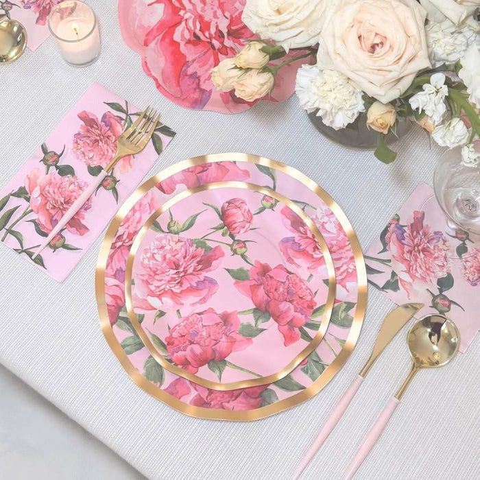 The Pink Peonies Table Setting by Sophistiplate LLC features designer floral plates, matching napkins, gold-pink cutlery, a wine glass, a candle, and pink-white flowers on a light tablecloth.