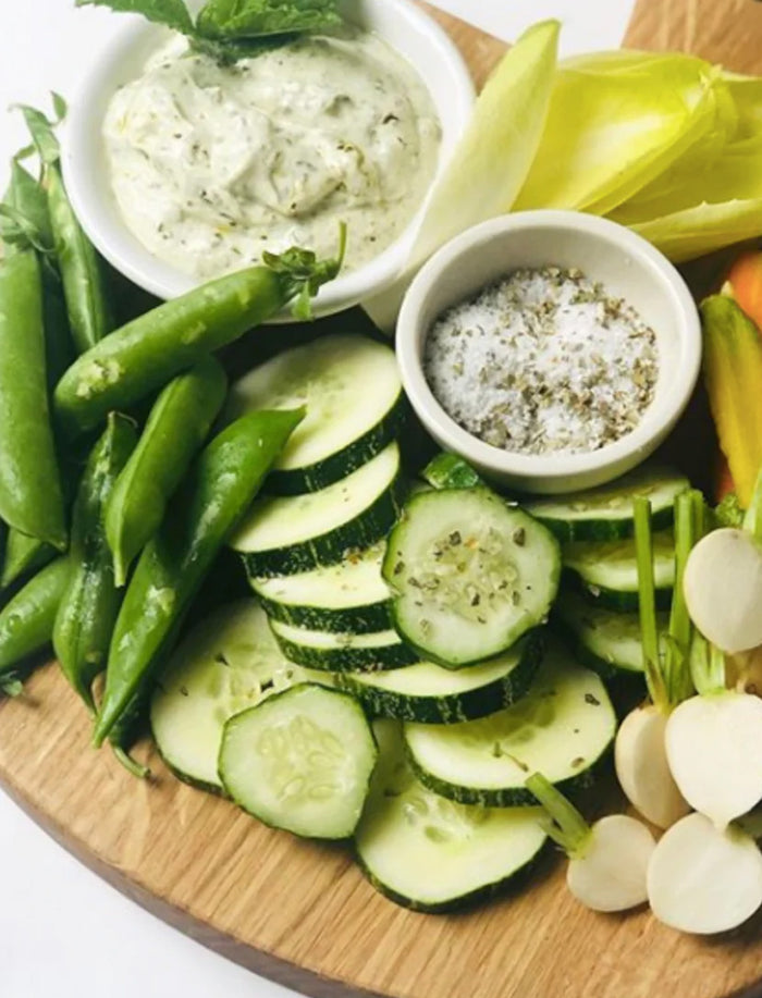 A wooden charcuterie board with sliced cucumbers, snap peas, endive, radishes, creamy herb dip, and a Bella Cucina Savory Salt Duo Gift Set for gourmet seasoning.