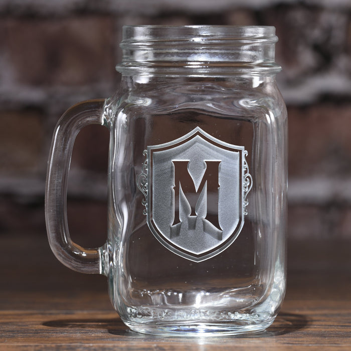 Mason jar mug sand-carved with the initial "M" on an ornate shield, against a brick wall.