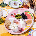 The Summer Soirée Edit by Maple features a pink melamine plate with bread, cheese, olives, blackberries, marshmallows, and watermelon on a yellow plaid cloth, complete with glassware and a picnic basket nearby.