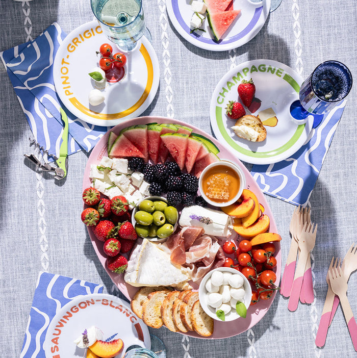 The Summer Soirée Edit by Maple features a vibrant picnic with a charcuterie tray of cheese, berries, prosciutto, olives, honey, peaches, bread & mozzarella on a blue tablecloth, surrounded by melamine plates of fruit and drinks.