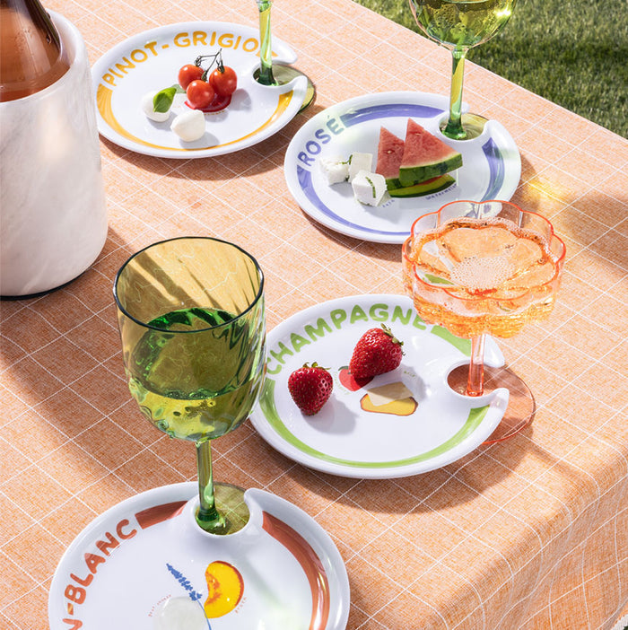 Maple’s The Summer Soirée Edit features a picnic table with a peach plaid cloth, wine glasses, and melamine plates labeled by wine type. Each plate has fruit or cheese and a notch to hold your glass—perfect for stylish outdoor gatherings.