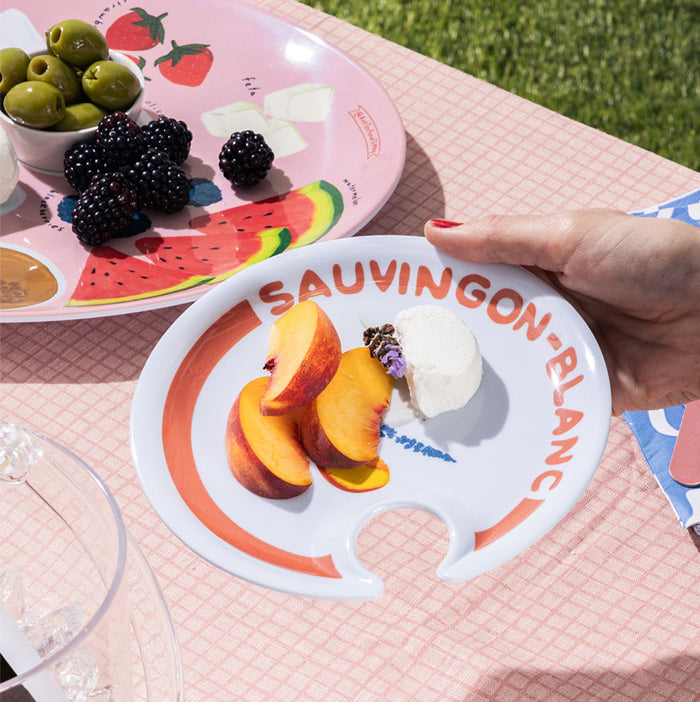 A hand holds Maple’s melamine plate from The Summer Soirée Edit with peach slices, cheese, and lavender. In the background, another tray showcases olives, blackberries, cheese, and watermelon.