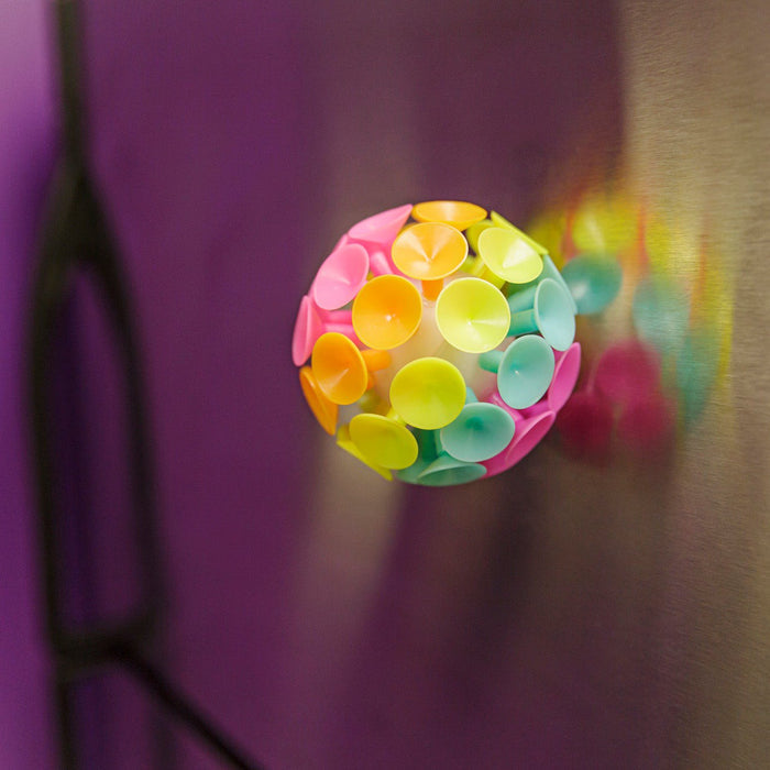 A Toysmith Jumbo Suction Ball, featuring overlapping pink, yellow, green, and blue discs, sticks to a metallic surface and casts a vibrant reflection. This fun sensory toy by Toysmith is perfect for kids ages 3+. A blurred purple background is visible.