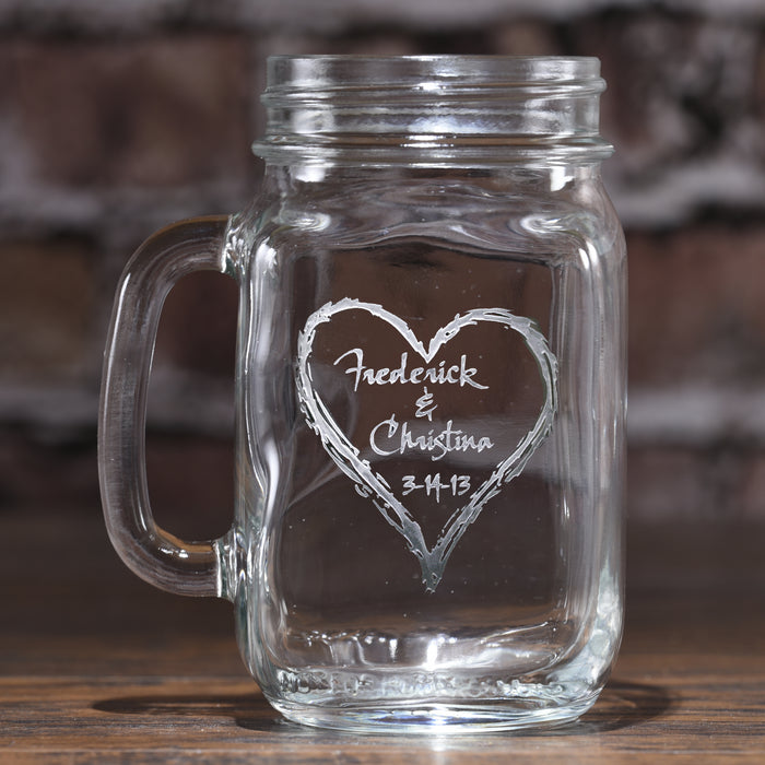 Mason jar mug sand-carved with the names "Frederick & Christina" and a date inside a hand-drawn heart, against a brick wall.